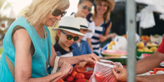 Resident from Apple Valley Villa shopping at a local farmers market, selecting fresh produce with children and families in the background.